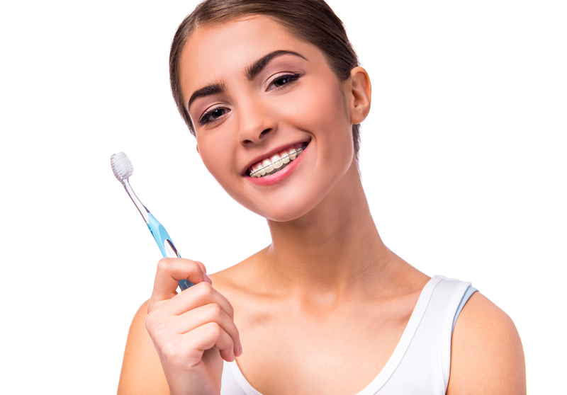 A brunette attractive teen girl that is smiling with her ceramic braces on her teeth and holding up a toothbrush.