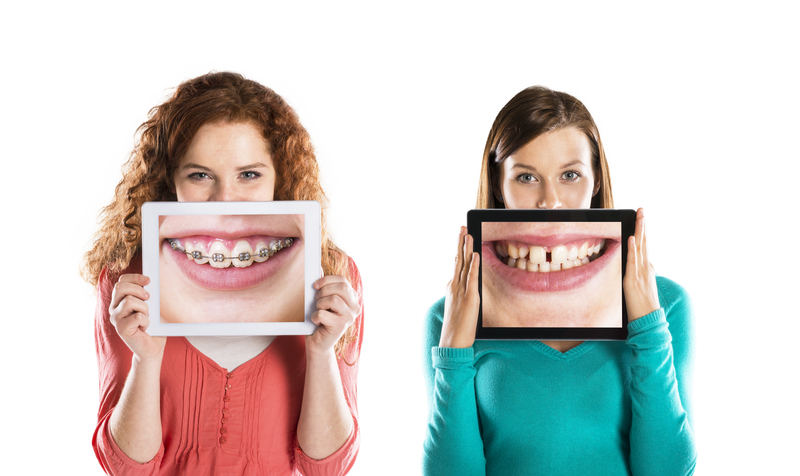 Two girls standing side-by-side that are both holding up photos in front of their face. One photo shows teeth that are crooked and the other shows teeth that are getting straightened with braces.
