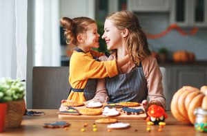 A mother and young daughter smiling as they start to hug. They are sitting in front of Halloween sugar cookies they just finished frosting.