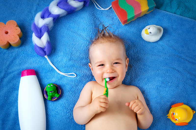 Image of a smiling infant with a toothbrush. He is on a blue towel background and there are various bath items around him.