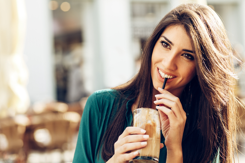 A beautiful brunette young adult woman that is sipping on a coffee drink. She is wearing green.