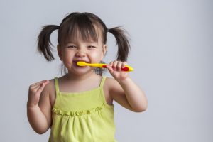 A young brunette toddler that is smiling and brushing her teeth.