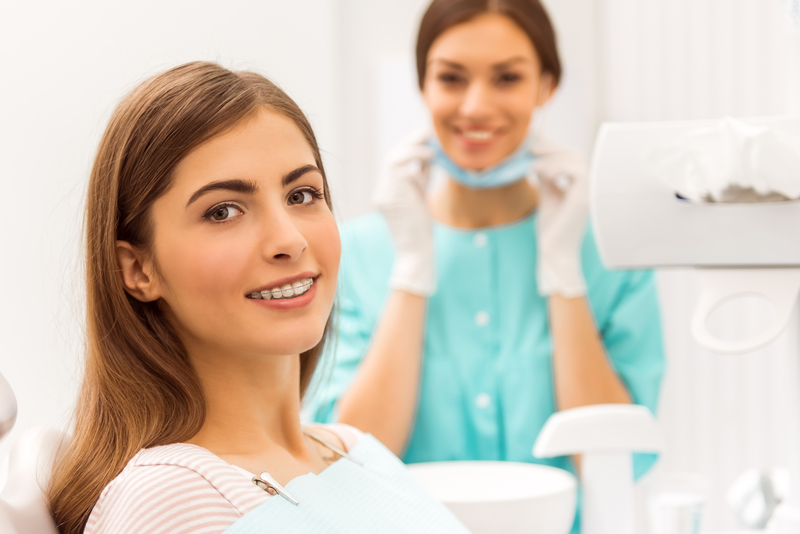 A young adult brunette woman with braces that is sitting in a dental office with a dental hygienist smiling in the background.