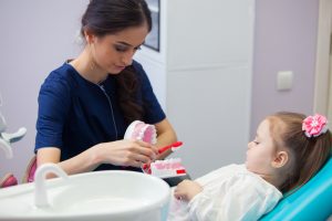 A dental hygienist in a pediatric office showing a young girl how to brush her teeth on a dental model.